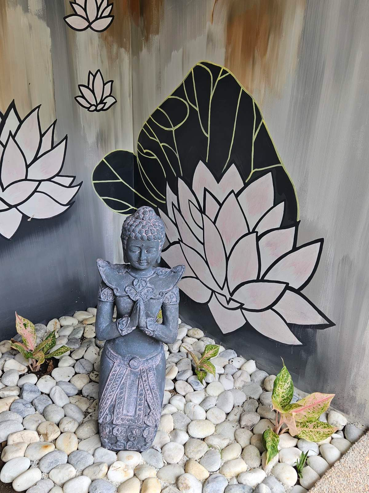 Small decorative statue among white stones and plants in front of a lotus mural.